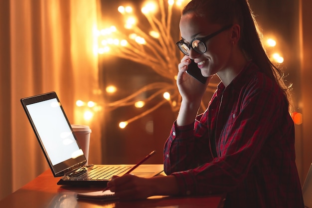 Concetto di affari, superlavoro, scadenza e persone. giovane donna con gli occhiali che lavora online usando laptop e telefono tardi a casa. lavoratore notturno e serale