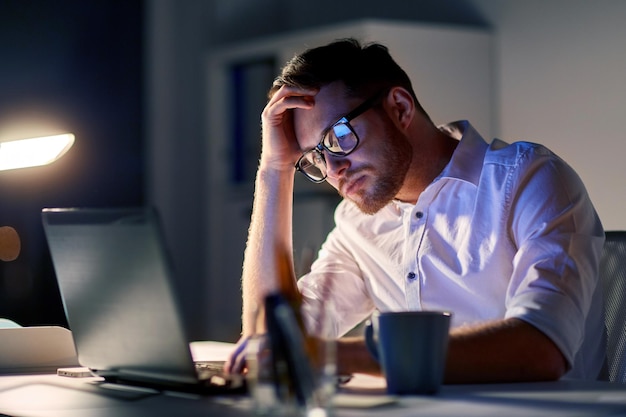 business, overwork, people, deadline and technology concept - stressed businessman in glasses with laptop computer thinking at night office
