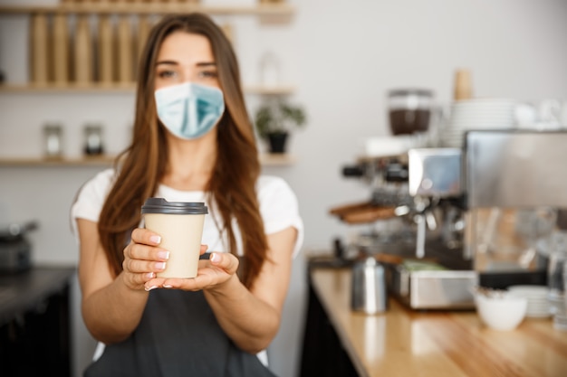 Business Owner Concept - Beautiful Caucasian Barista in face mask offers disposable take away hot coffee at the modern coffee shop