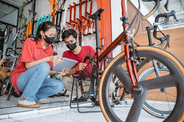 事業主とマスクの整備士がタブレットを使用している顧客のために新しい自転車部品をチェックします