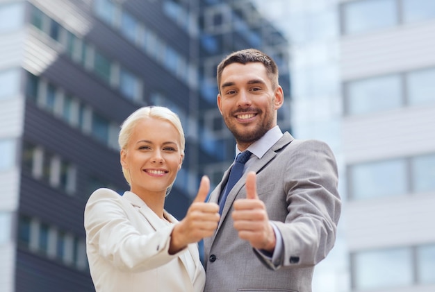 business, partnership, success, gesture and people concept - smiling businessman and businesswoman showing thumbs up over office building