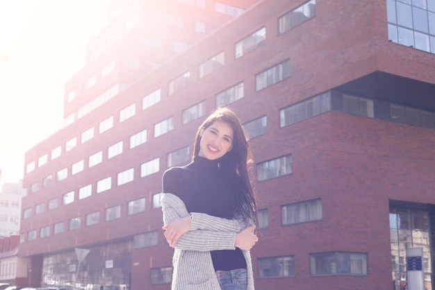 Concetto di affari, bellezza della gente e stile di vita - giovane donna d'affari bruna di moda contro il centro affari al tramonto. ragazza in una giacca nera e jeans blu. sorriso, professionalità. luce del sole