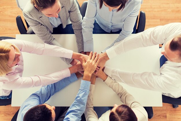 business, people, cooperation and team work concept - close up of creative team sitting at table and holding hands on top of each other in office