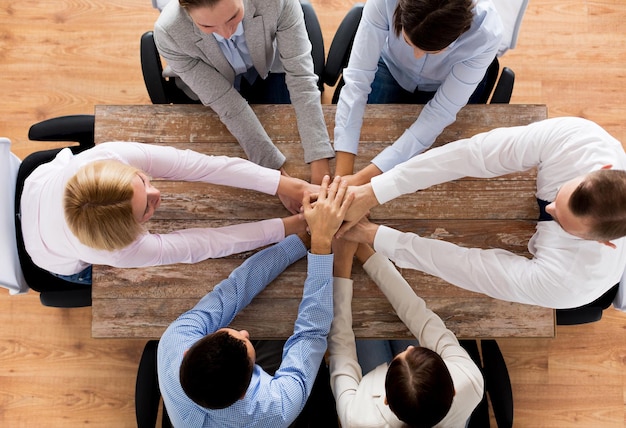 концепция бизнеса, людей, сотрудничества и командной работы - крупный план творческой команды, сидящей за столом и держащейся за руки друг над другом в офисе