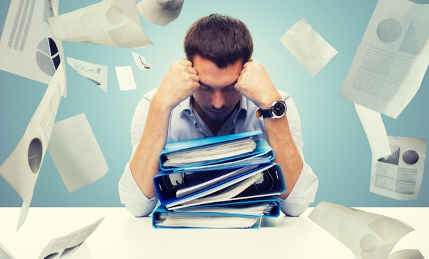 концепция бизнеса, людей, крайнего срока, стресса и работы с документами - грустный бизнесмен со стопкой папок и падающими бумагами на синем фоне