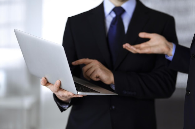 Business people discussing a presentation at meeting, standing in a modern office. Unknown businessman with a colleague search for some information at the laptop, lawyers at negotiation. Teamwork and