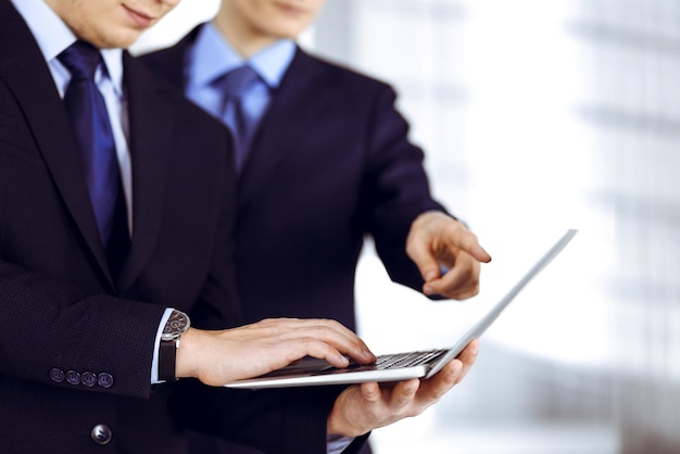 Business people discussing a presentation at meeting, standing in a modern office. Unknown businessman with a colleague search for some information at the laptop, lawyers at negotiation. Teamwork and