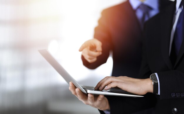 Business people discussing a presentation at meeting, standing in a sunny modern office. Unknown businessman with a colleague search for some information at the laptop, lawyers at negotiation. Teamwor