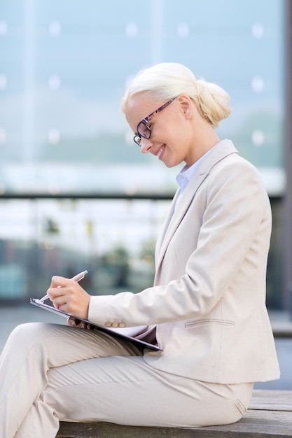 business, people and education concept - young smiling businesswoman in glasses with notepad over office building