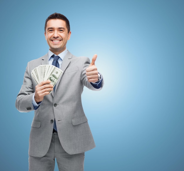 Business, people and finances concept - smiling businessman with dollar money showing thumbs up over blue background