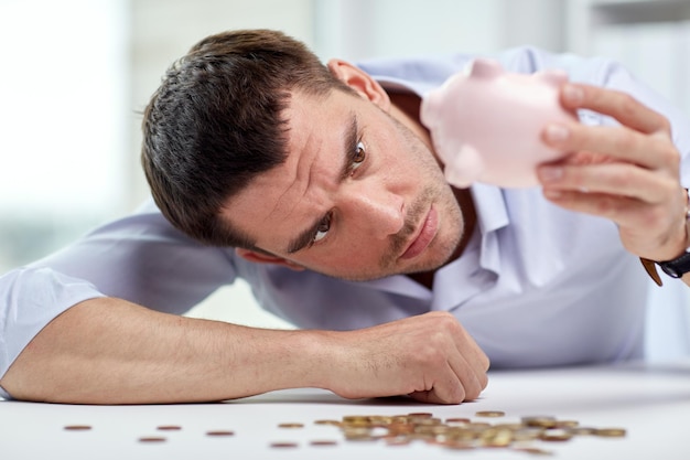 business, people, finances, crisis and money saving concept - businessman shaking piggy bank and getting coins at office