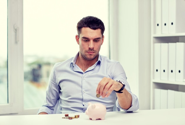 business, people, finances and money saving concept - businessman with piggy bank and coins at office