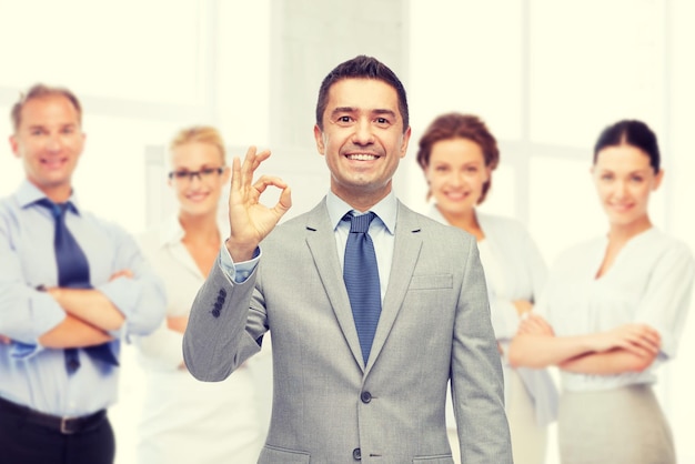 business, people, gesture and success concept - happy smiling businessman in suit with team over office room background showing ok hand sign