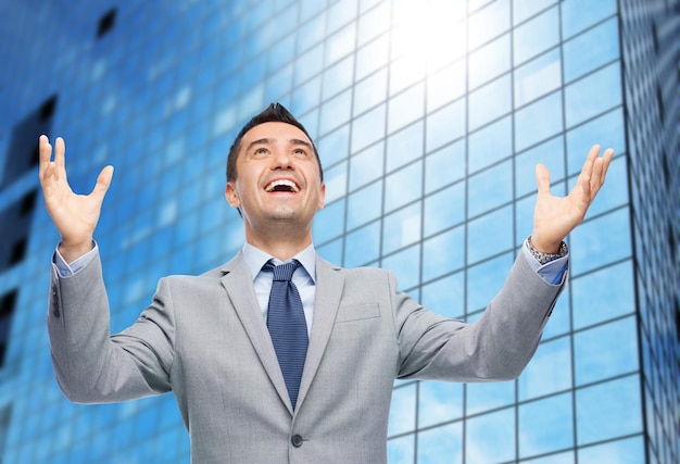 business, people and happiness concept - happy businessman in suit with raised hands laughing and looking up over office building background