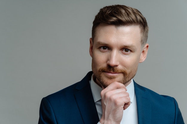 Business people. Headshot of handsome unshaved businessman or male entrepreneur in blue navy suit touching his chin and looking at camera thoughtfully while posing against grey studio background