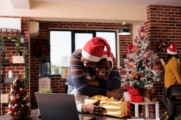 Business people hugging after exchanging christmas gifts in office decorated with festive ornaments and xmas tree. Colleagues celebrating winter holiday in workplace with seasonal decorations.