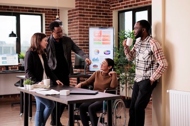 Business people meeting on break in disability friendly office, laughing with impaired asian woman. Female wheelchair user with physical health condition chatting with work colleagues.