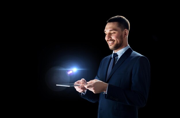 Business, people and modern technology concept - smiling businessman in suit working with transparent tablet pc computer over black background