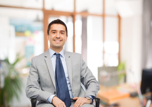 business, people and office concept - happy businessman in suit sitting in chair over office room background