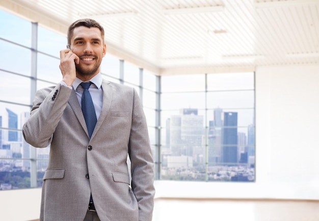 business, people and office concept - happy young businessman calling on smartphone over office room or new apartment background