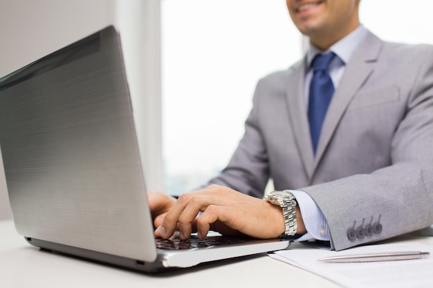 концепция бизнеса, людей, документов и технологий - крупный план улыбающегося бизнесмена с ноутбуком и бумагами, работающего в офисе