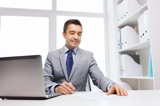 концепция бизнеса, людей, документов и технологий - улыбающийся бизнесмен с ноутбуком и бумагами, работающий в офисе