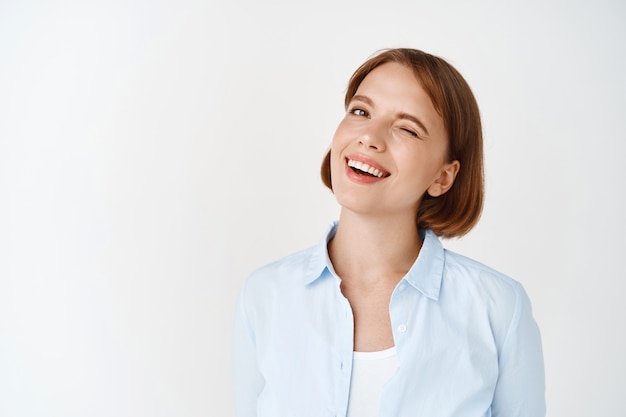Business people. Portrait of young woman with short hair, winking and smiling motivated, standing upbeat on white wall