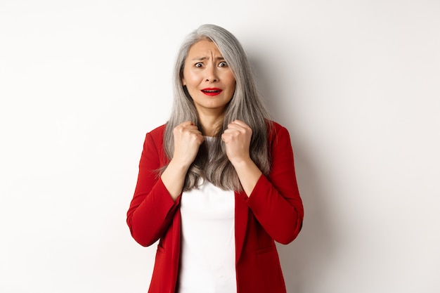 Business people. Scared mature asian woman looking terrified, trembling from fear, standing in red blazer over white background