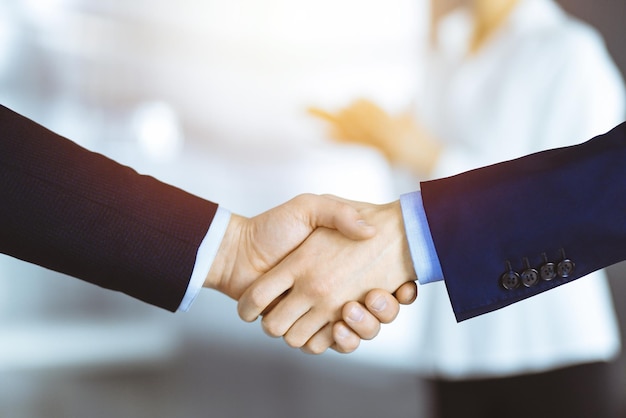 Business people shaking hands at meeting or negotiation, close-up. Group of unknown businessmen and a woman standing in a sunny modern office. Teamwork, partnership and handshake concept