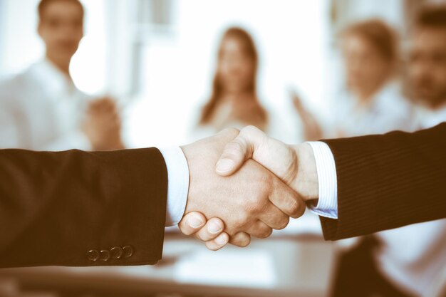 Business people shaking hands at meeting while theirs colleagues clapping and applausing. Group of unknown businessmen and women in modern white office. Success teamwork, partnership and handshake con