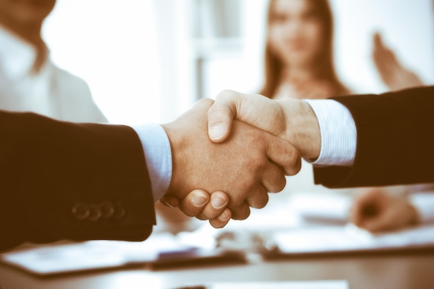 Business people shaking hands at meeting while theirs colleagues clapping and applausing. Group of unknown businessmen and women in modern white office. Success teamwork, partnership and handshake con