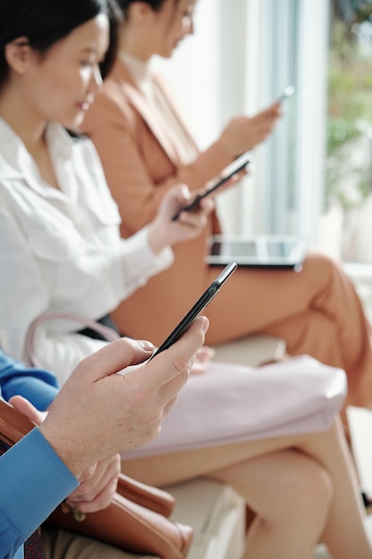 Business people sitting on chairs when waiting in queue and reading articles or checking social media on smartphones