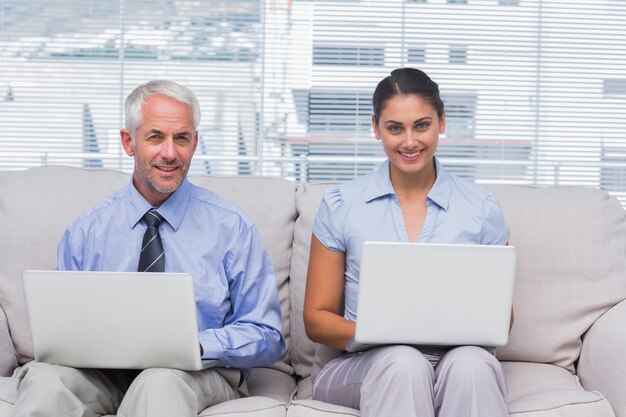 Business people sitting on sofa using their laptops and smiling at camera