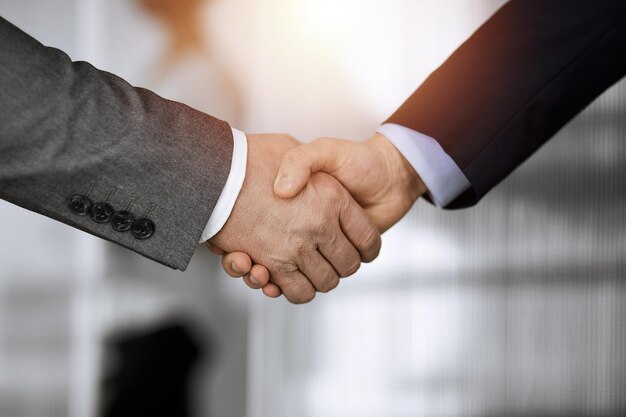 日当たりの良いオフィスで立って握手をするビジネスマン、接写。握手とマーケティング。