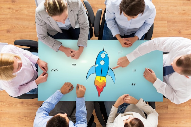 business, people, startup, development and team work concept - close up of creative team sitting at table and pointing finger to rocket icon in office