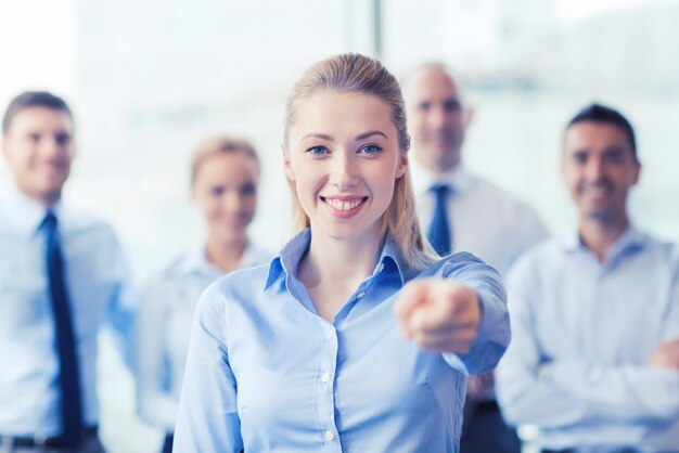 Business, people and teamwork concept - smiling businesswoman pointing finger on you with group of businesspeople in office