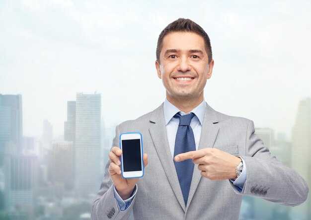 business, people and technology concept - happy smiling businessman in suit showing smartphone black blank screen over city background