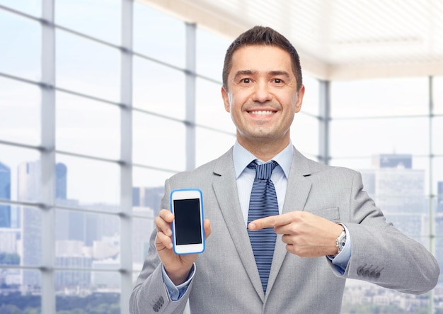 business, people and technology concept - happy smiling businessman in suit showing smartphone black blank screen over city office window background