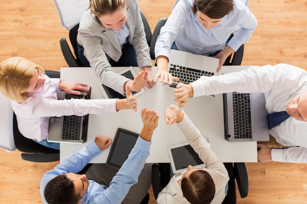 business, people, technology and team work concept - close up of creative team with laptop and tablet pc computers showing high five gesture and sitting at table in office