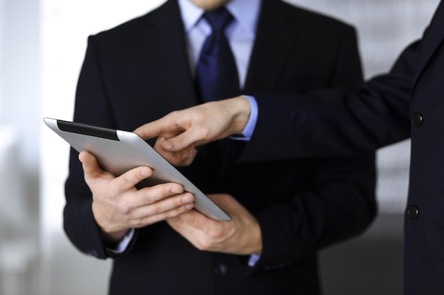 Business people use a tablet computer for discussion of their new project, standing in a modern office. Unknown businessman or male entrepreneur with a colleague at workplace. Teamwork and partnership