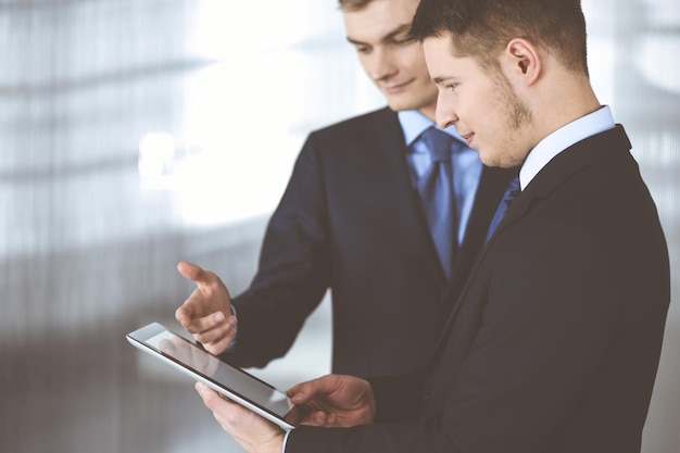 Business people, using a tablet computer, while standing in a modern office. Unknown businessman with a colleague at workplace. Teamwork and partnership concept.