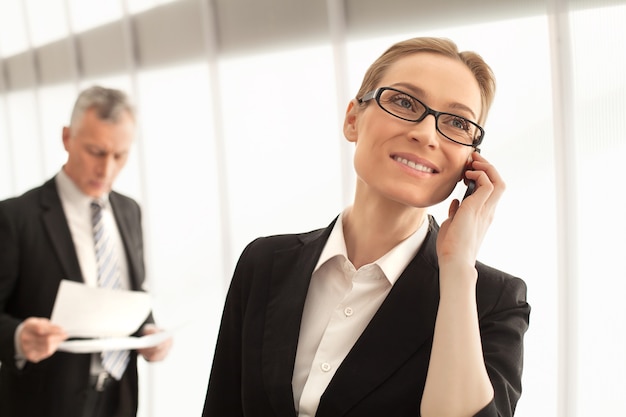 Foto uomini d'affari al lavoro. bella donna d'affari matura che parla al telefono cellulare mentre l'uomo in abiti da cerimonia in piedi sullo sfondo