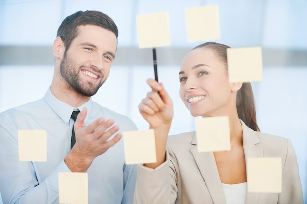 Business planning. Two cheerful business people discussing something while pointing adhesive notes attached to the glass