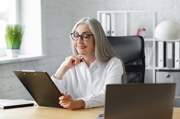 Business portrait of beautiful smiling elderly woman wearing eyeglasses sitting at workplace. Grey-haired senior businessman working in office with tables, charts, reports.