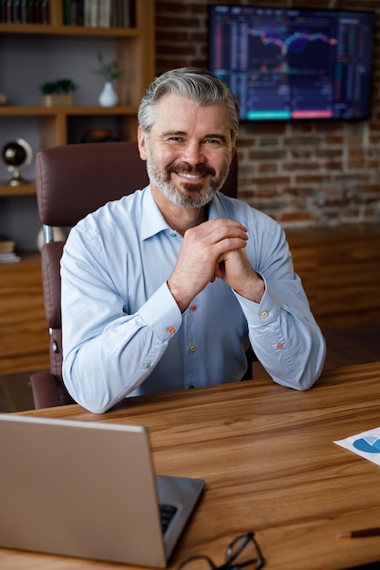 Business portrait of confident handsome bearded businessman looking at camera and smiling Front close up portrait of Caucasian grayhaired man indoors at home office Human face