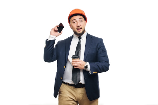 Business portrait of surprised contractor and developer, talking about phone. Businessman in hard hat with cup of coffee standing over white background. News and coffee brake concept