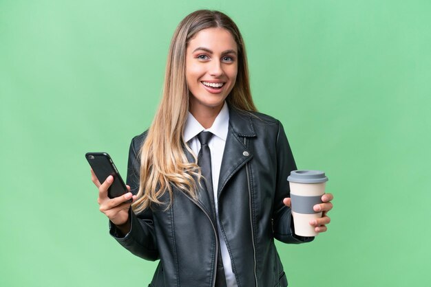 Business pretty Uruguayan woman wearing a biker jacket over isolated background holding coffee to take away and a mobile