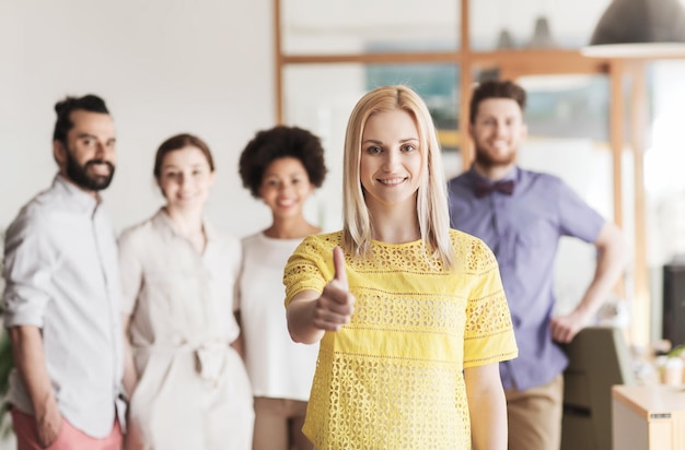 business, startup, people, gesture and teamwork concept - happy young woman showing thumbs up over creative team in office