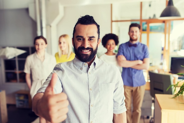 business, startup, people, success and teamwork concept - happy young man with beard over creative team showing thumbs up in office