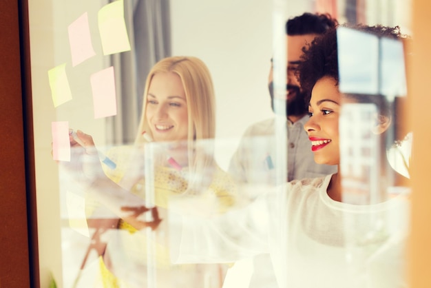 비즈니스, 시작, 계획, 관리 및 사람 개념 - 사무실 유리판에 스티커를 붙이는 행복한 국제 크리에이티브 팀